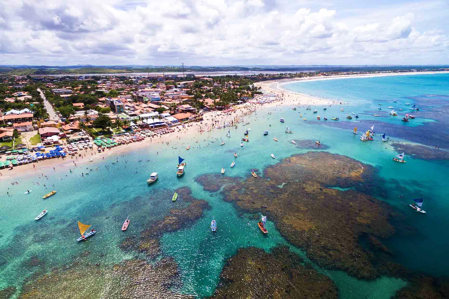 Aerial view of a crowded Brazilian beach with turquoise waters, moored boats, and colorful residential buildings lining the s